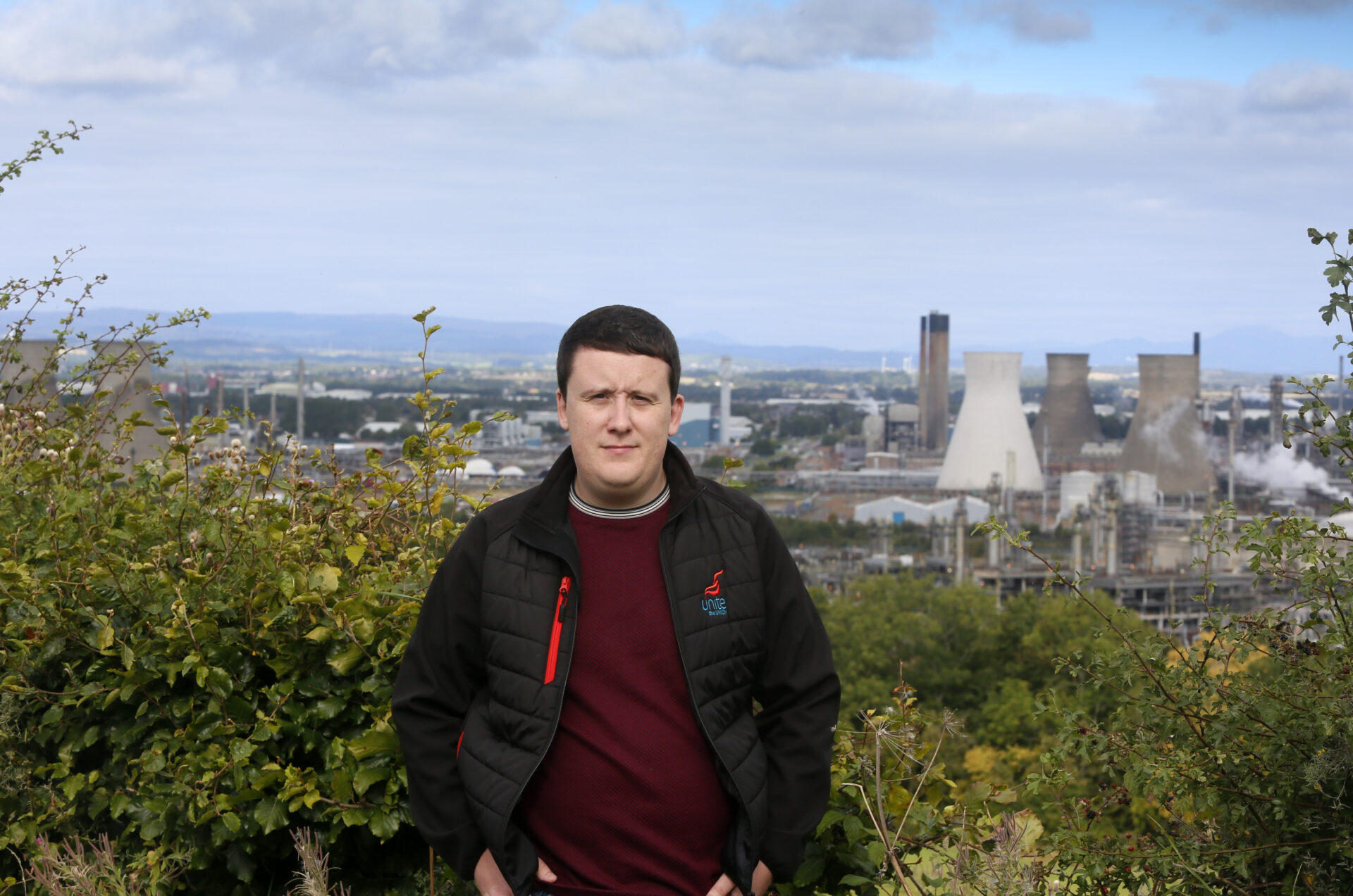 Unite union representative Chris Hamilton standing in a grassy field with Grangemouth refinery’s cooling towers and chimneys in the background.