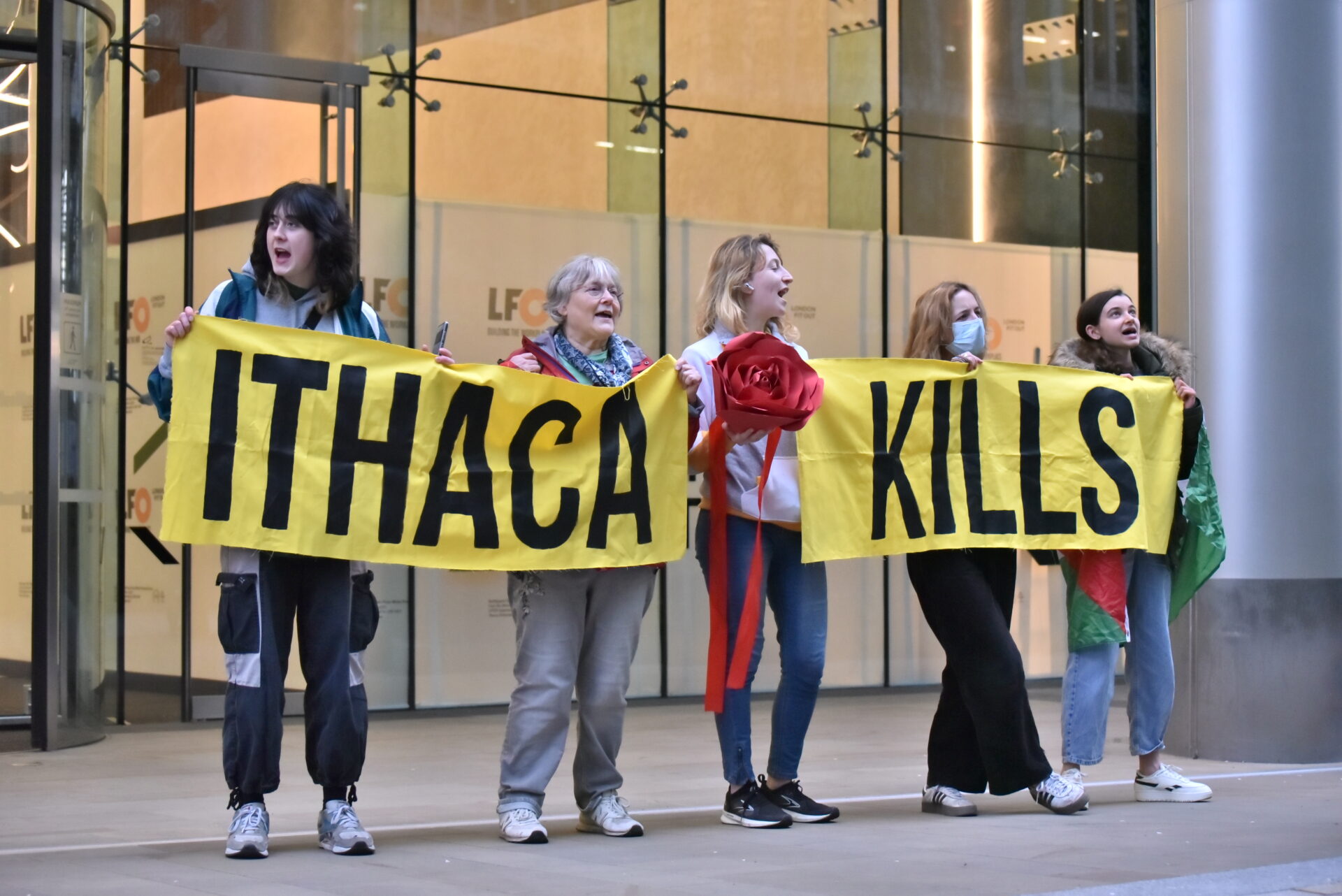 Protesters stand in front of glass offices holding a yellow banner that says “ITHACA KILLS.” Some wear Palestinian flags or carry props, with one person holding a large red paper flower.