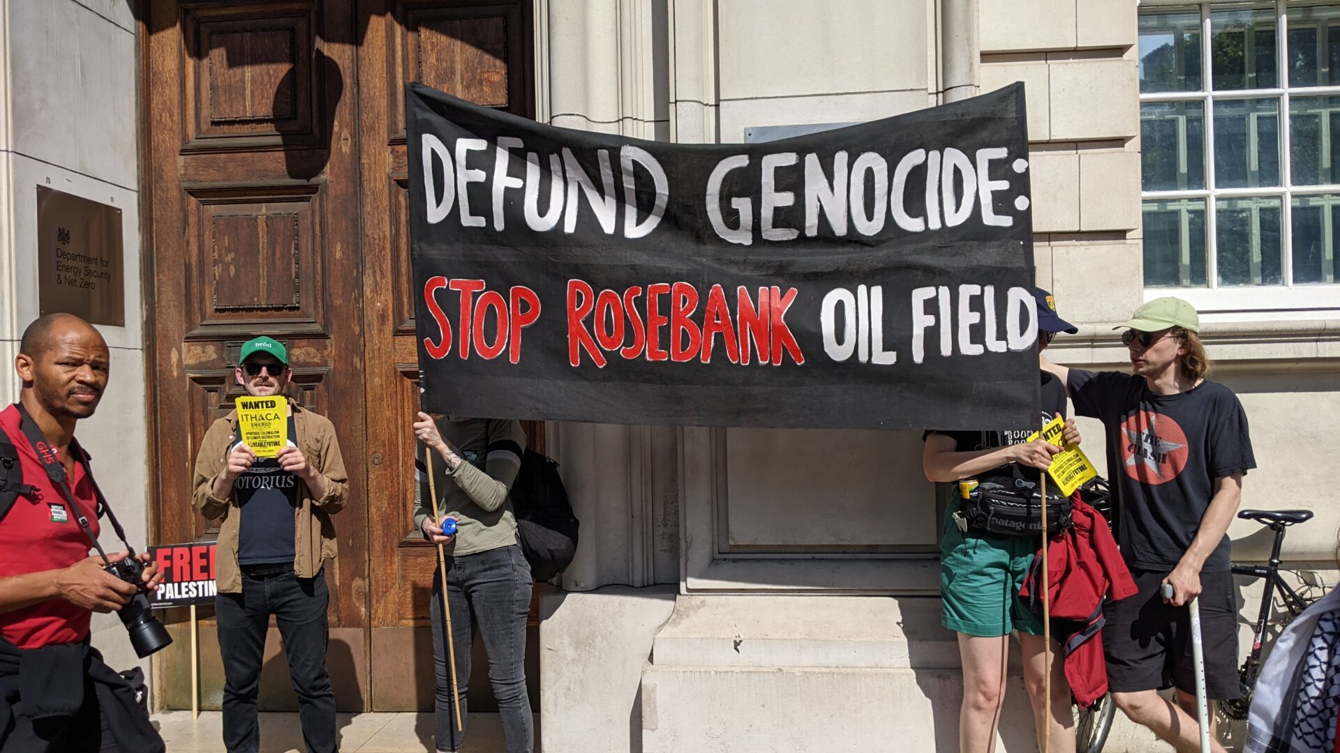 Protesters outside the UK Department for Energy hold a large black banner reading “DEFUND GENOCIDE: STOP ROSEBANK OIL FIELD.” Others hold smaller yellow “Wanted Ithaca Energy” signs.
