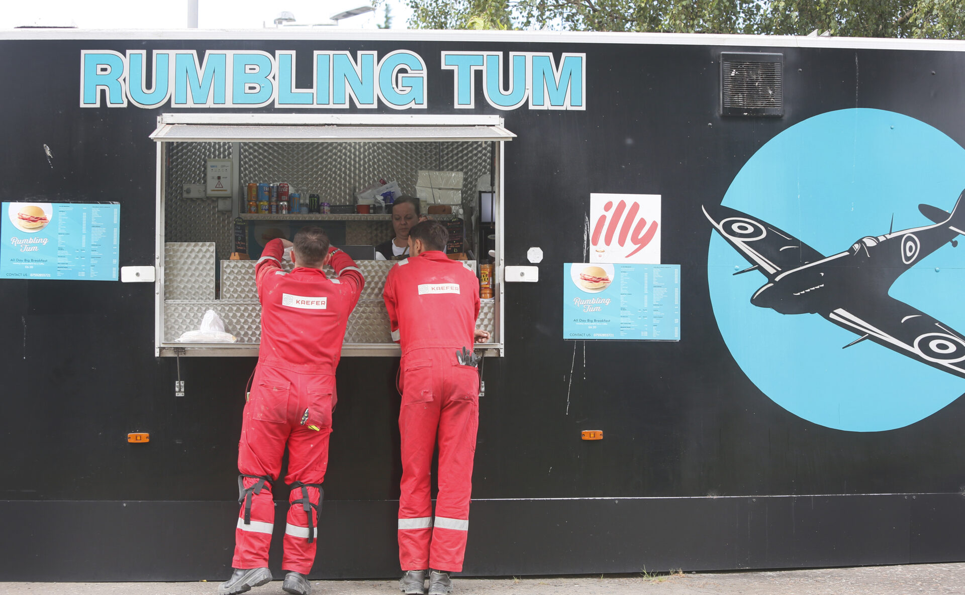 Two refinery contractors in red overalls order food at a black van with “Rumbling Tum” written above the serving hatch.
