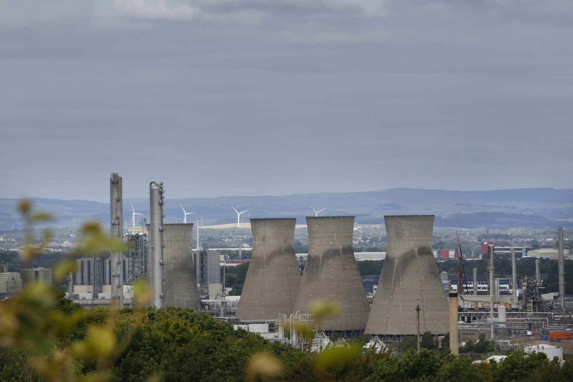 Article headline: Power struggle: The billionaire, the Chinese oil giant and Grangemouth's forgotten workers Image description: Cooling towers and chimneys of the Grangemouth oil refinery rising above the town, with wind turbines visible in the background.