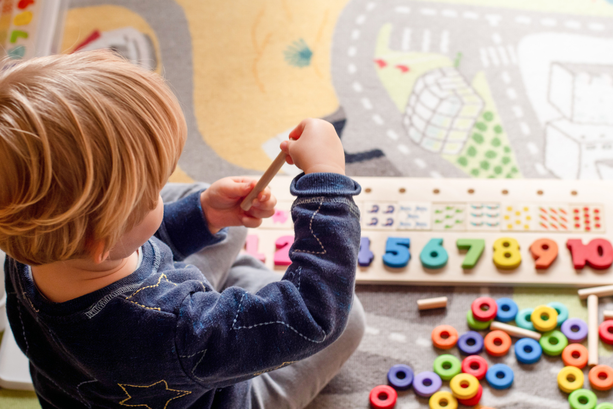 Young child sitting on a colourful play mat, playing with a wooden educational toy featuring numbers, pegs, and brightly coloured rings. The child is wearing a dark blue sweater with star patterns.