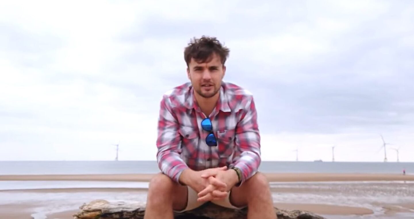 A man in a checked shirt and khaki shorts sits on driftwood at a beach, speaking directly to camera. Wind turbines are visible on the horizon behind him, under a grey sky.
