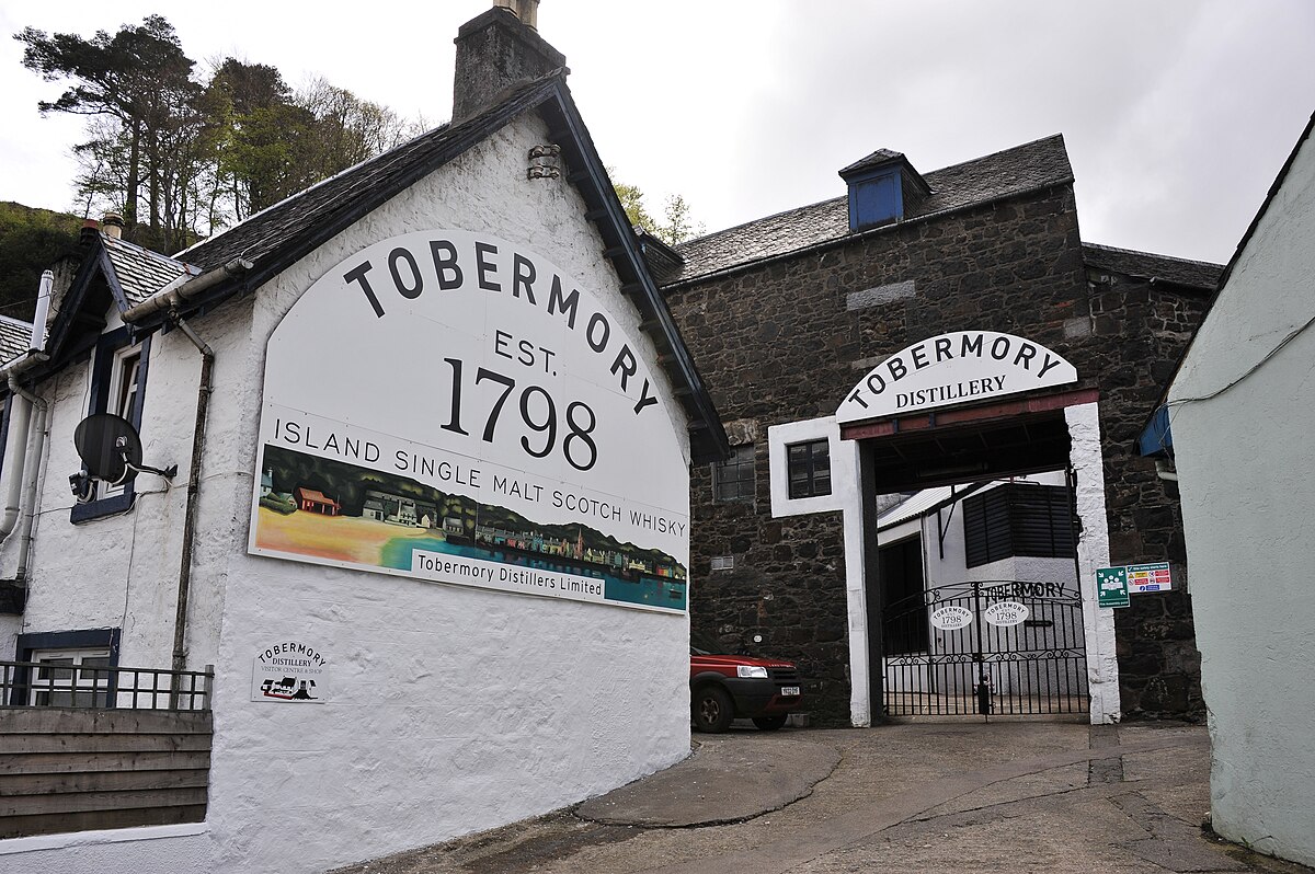 Image description: The image shows the Tobermory Distillery on the Isle of Mull. The distillery features traditional stone and whitewashed buildings, with a prominent sign reading "Tobermory Est. 1798 Island Single Malt Scotch Whisky." The entrance to the distillery is marked by an archway with "Tobermory Distillery" written above it, leading to a courtyard.