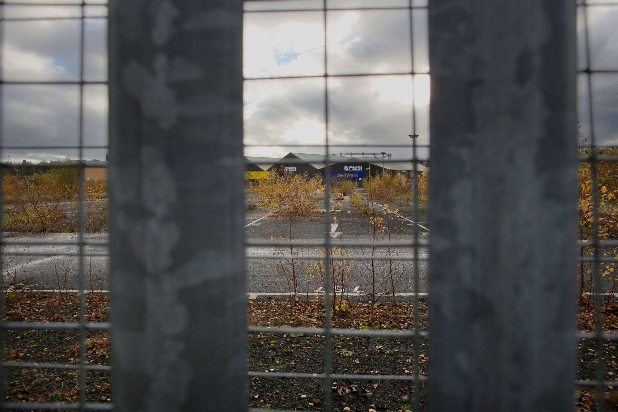 View through a wire fence showing an abandoned parking area with weeds growing through cracks and industrial buildings in the distance.