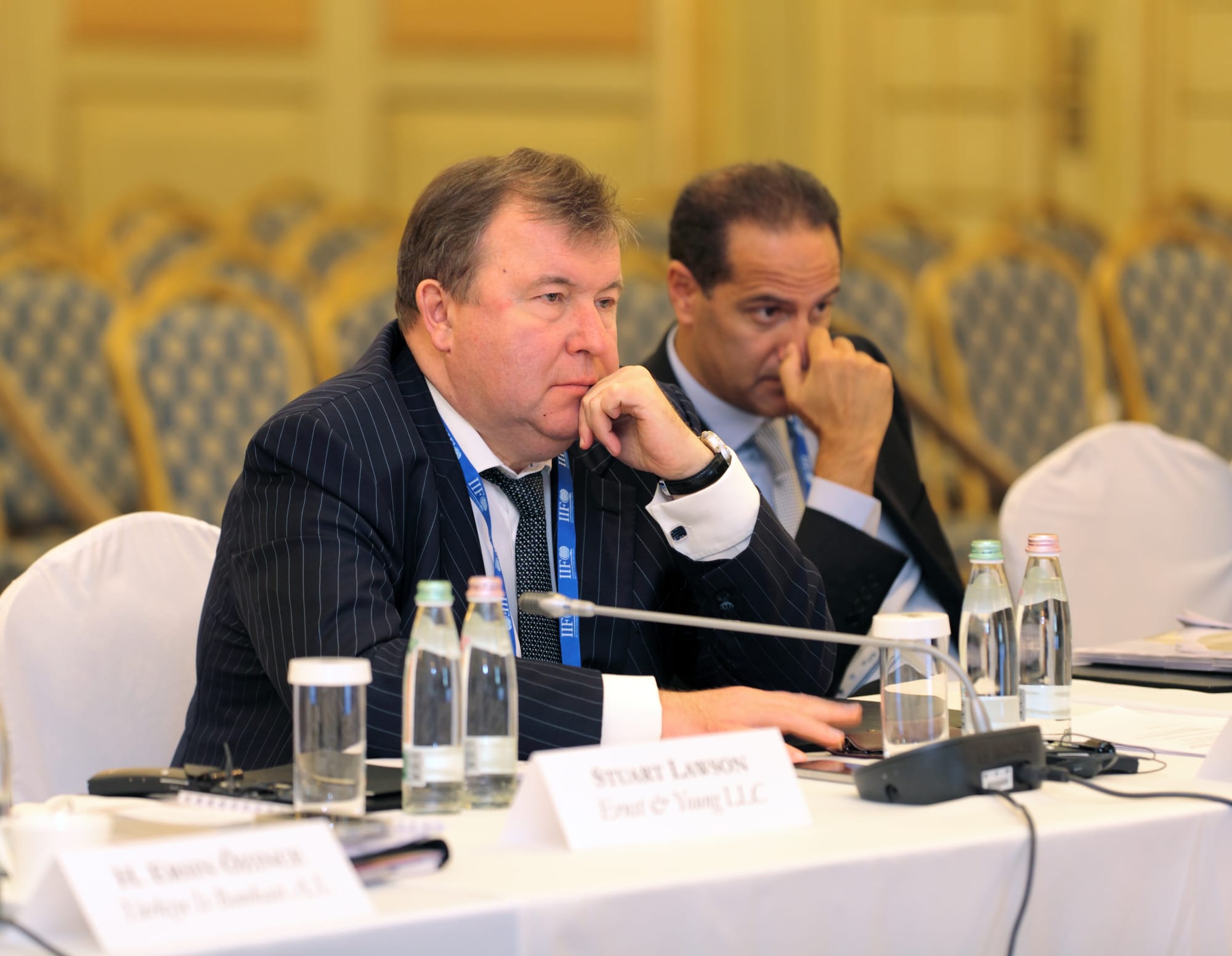 A man in a dark pinstripe suit sits at a conference table beside another attendee, with bottled water, microphones, and nameplates visible in front of them.