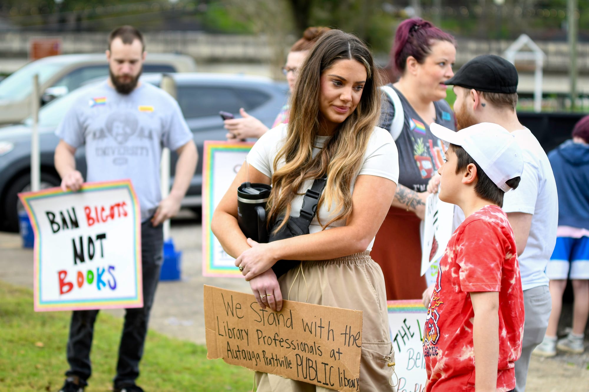 A group of adults and children stand at a peaceful outdoor protest. A woman holds a cardboard sign while speaking to a child. Behind them, other protesters hold signs, including one that reads “Ban bigots not books.”