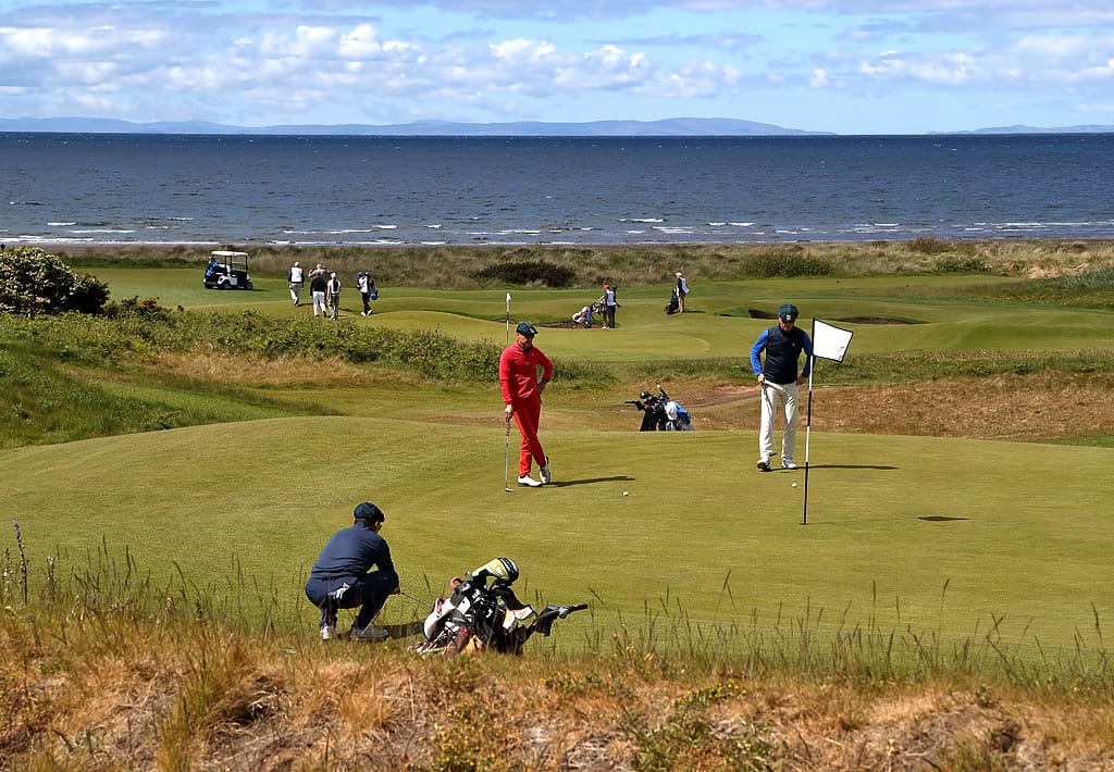 Golfers playing on a links course by the coast, with putting greens, golf bags and a buggy visible, and the sea in the background.