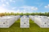 A field of large white battery storage units arranged in rows on green grass, with wind turbines and trees in the background under a blue sky.
