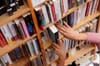 A close-up of a person’s hands removing a book from a wooden bookshelf filled with tightly packed books in a library.
