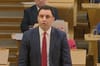 Anas Sarwar, leader of Scottish Labour, stands speaking in the Scottish Parliament chamber, wearing a dark suit and red tie. Other MSPs sit behind him at desks, listening.