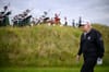 A man in golf attire walks across a grassy course while a group of bagpipers play in the background.